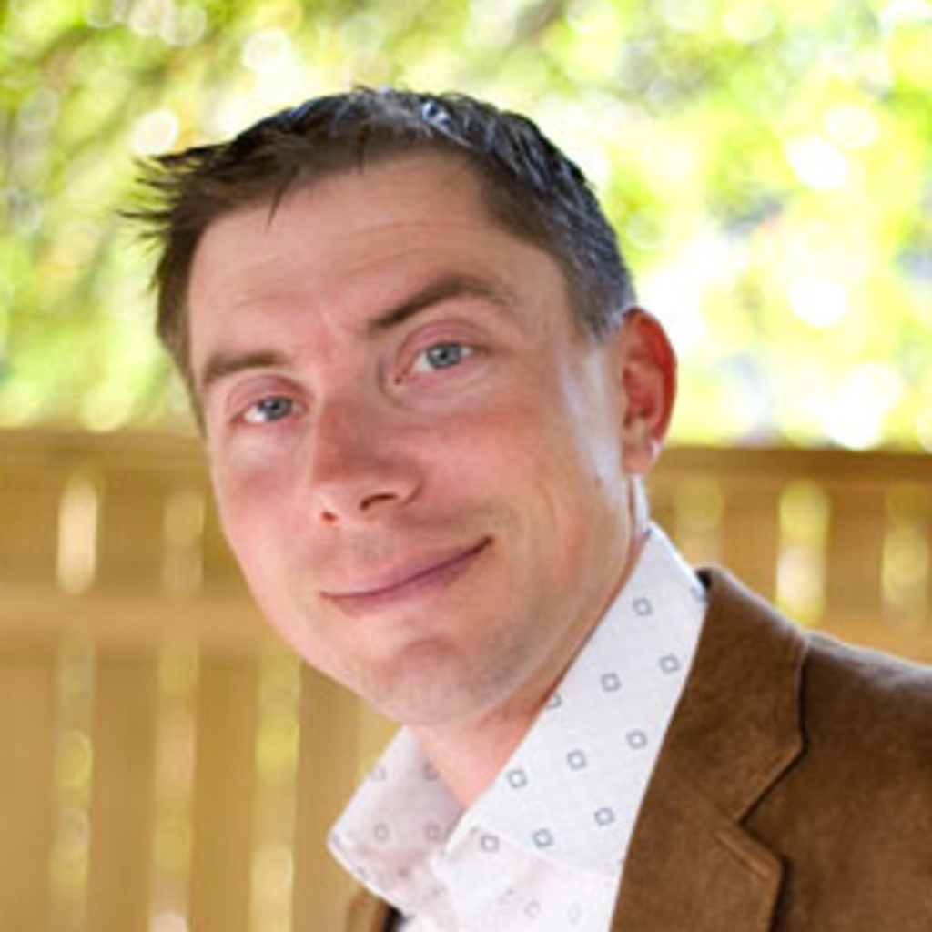 Matt Cohen smiles into the camera while wearing a brown suit jacket and white patterned button-up shirt.